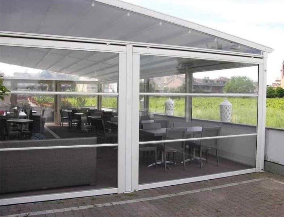 Una zona pranzo esterna bianca con pannelli in vetro, tavoli e sedie visibili all'interno, con uno sfondo urbano.
