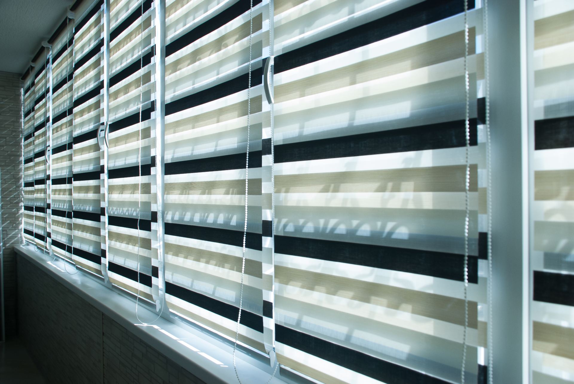 Fila di tende zebrate a doppio strato, con strisce alternate nere, beige e bianche, che coprono una finestra.