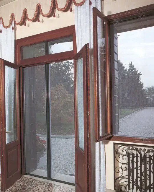 Porte e finestre in legno con zanzariere, che si aprono su un giardino, su una veranda beige chiaro.