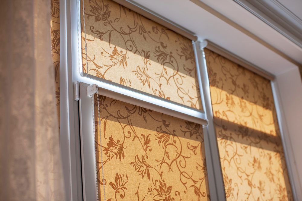 Primo piano di una finestra con tende a rullo beige con motivi floreali, parzialmente arrotolate, che proiettano ombre.