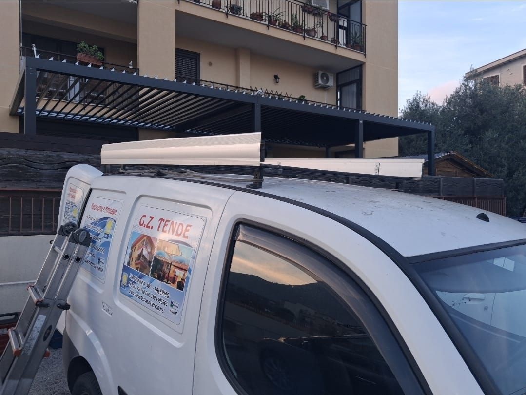 Furgone bianco con scala e pergolato nero sul tetto, parcheggiato vicino a un edificio.