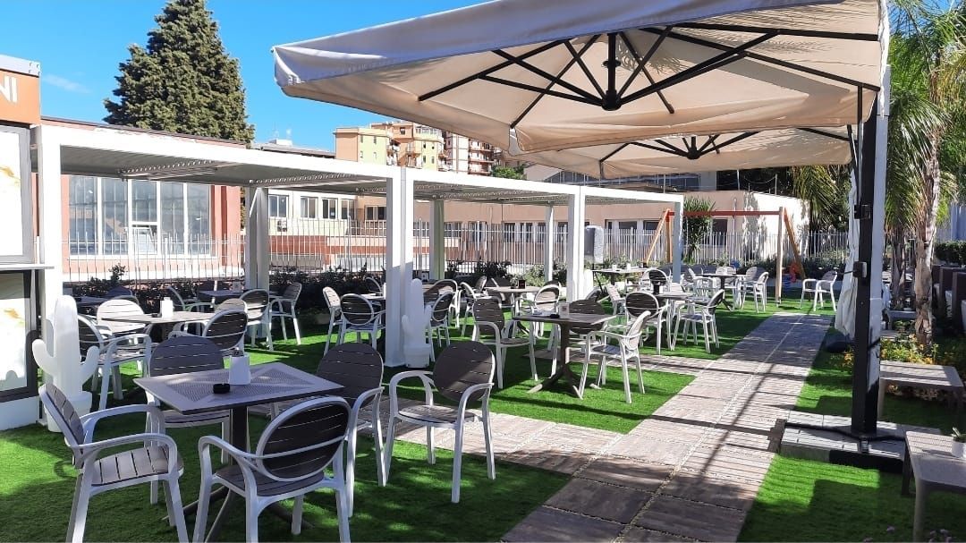 Bar all'aperto con tavoli, sedie, erba artificiale, ombrelloni e un edificio sullo sfondo.