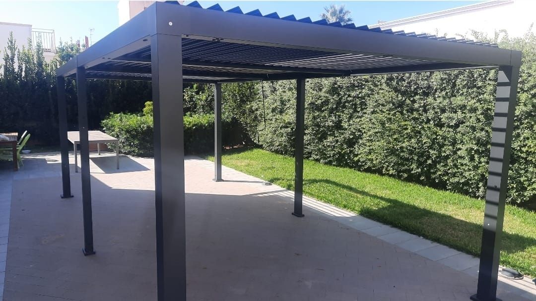 Pergola nera su patio in cemento, erba e cespugli verdi sullo sfondo.