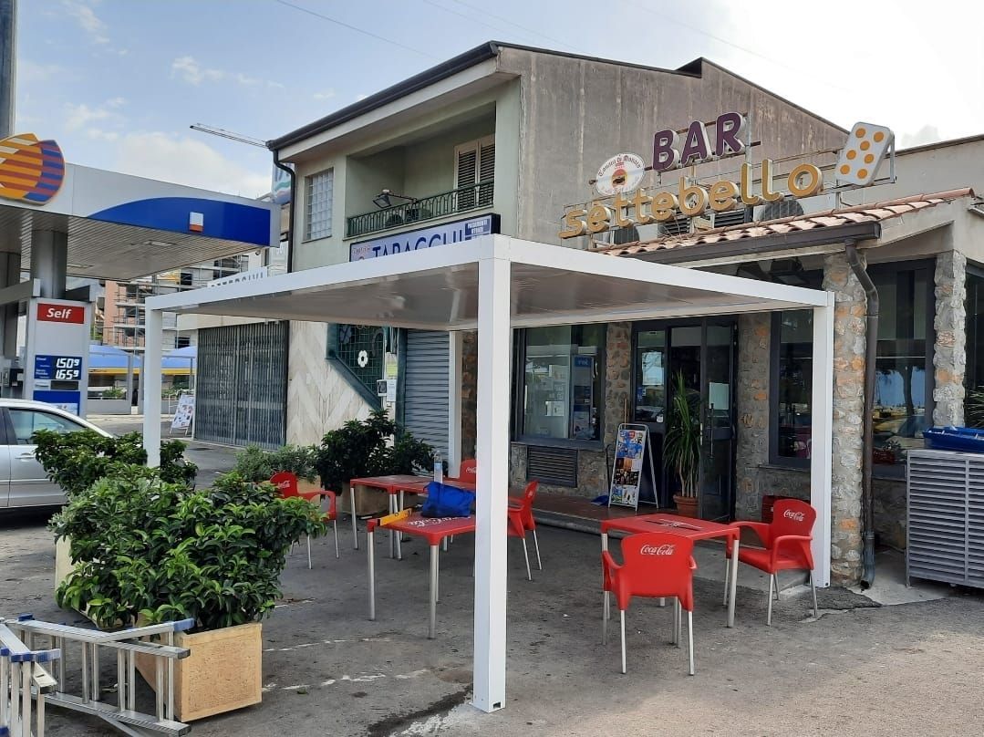 Bar all'aperto con tavoli e sedie rossi sotto un pergolato bianco, accanto a una stazione di servizio.