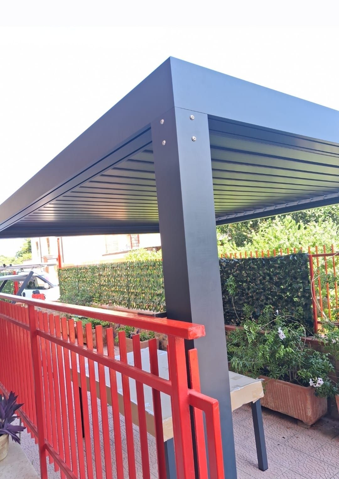 Pergola nera sopra la ringhiera rossa del patio. Siepe verde sullo sfondo.