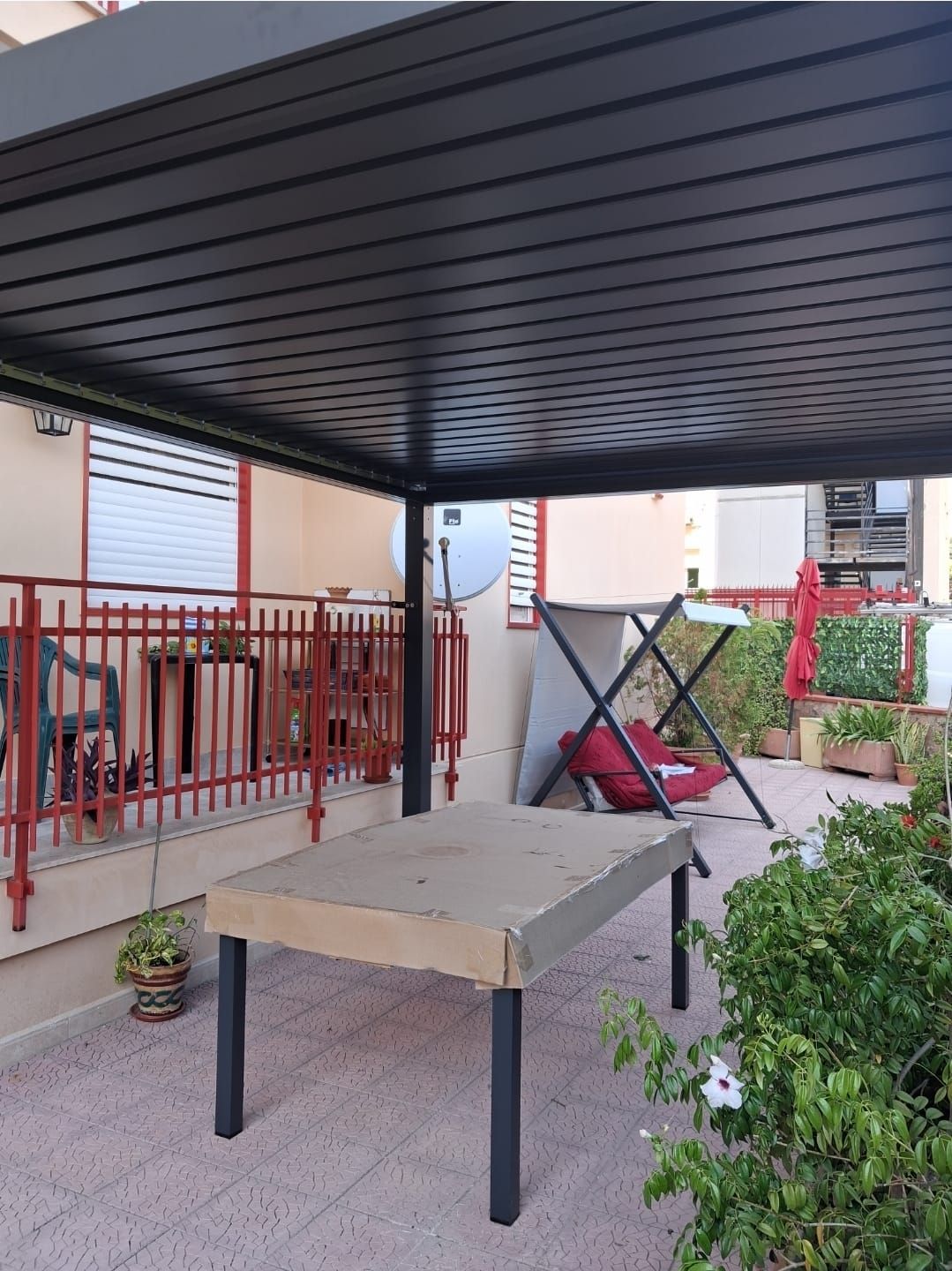 Patio esterno con pergolato nero, tavolo e ringhiera rossa.