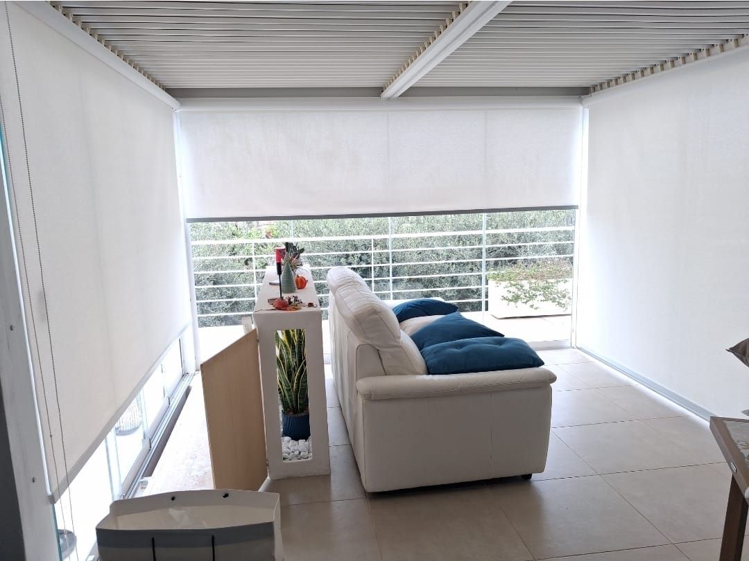 Patio soleggiato e recintato con mobili bianchi, divano e tende a rullo bianche. Balcone con vista sul verde.