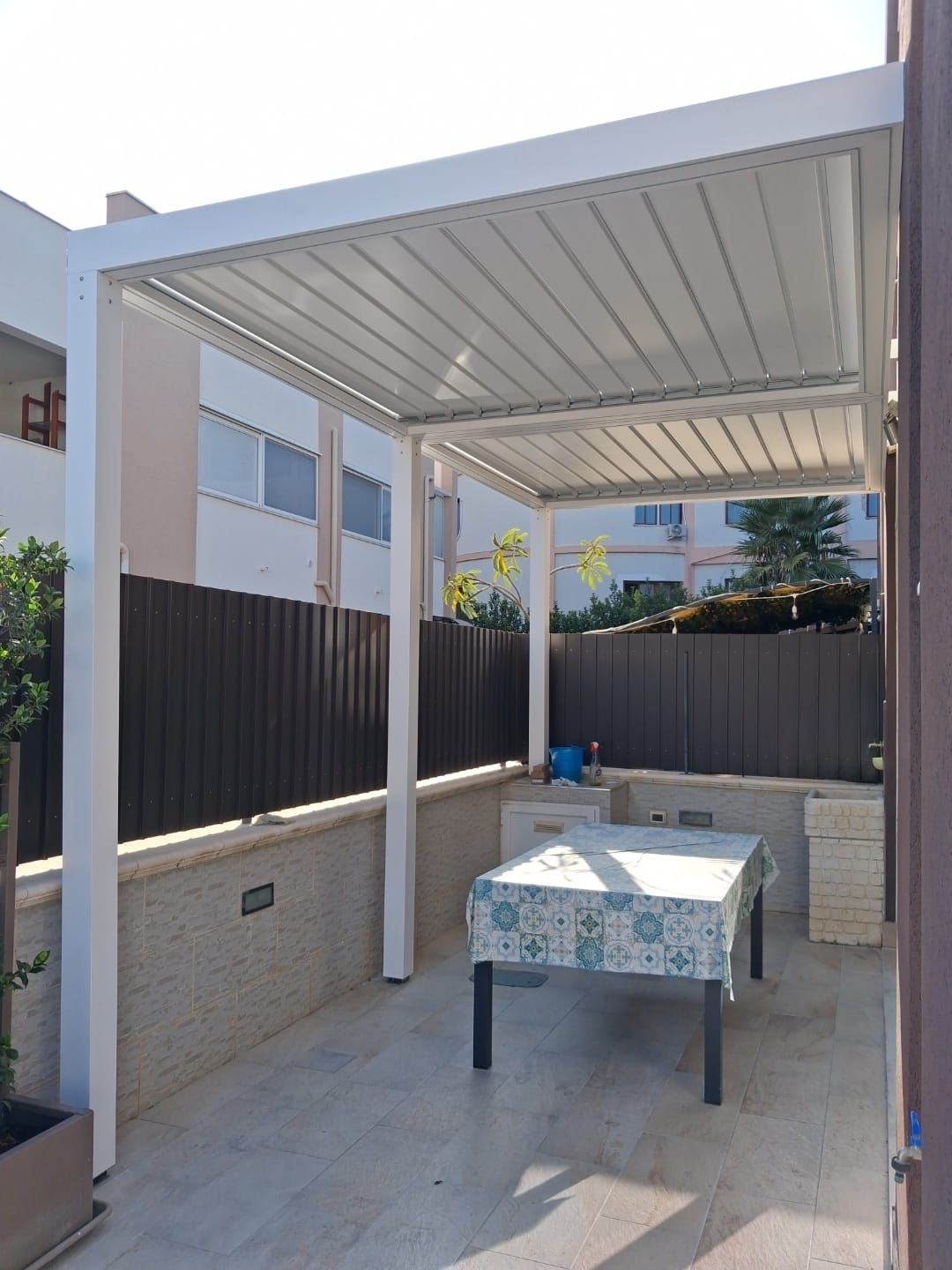 Pergola bianca sopra un patio con tavolo e sedie, adiacente a una recinzione e a un edificio.