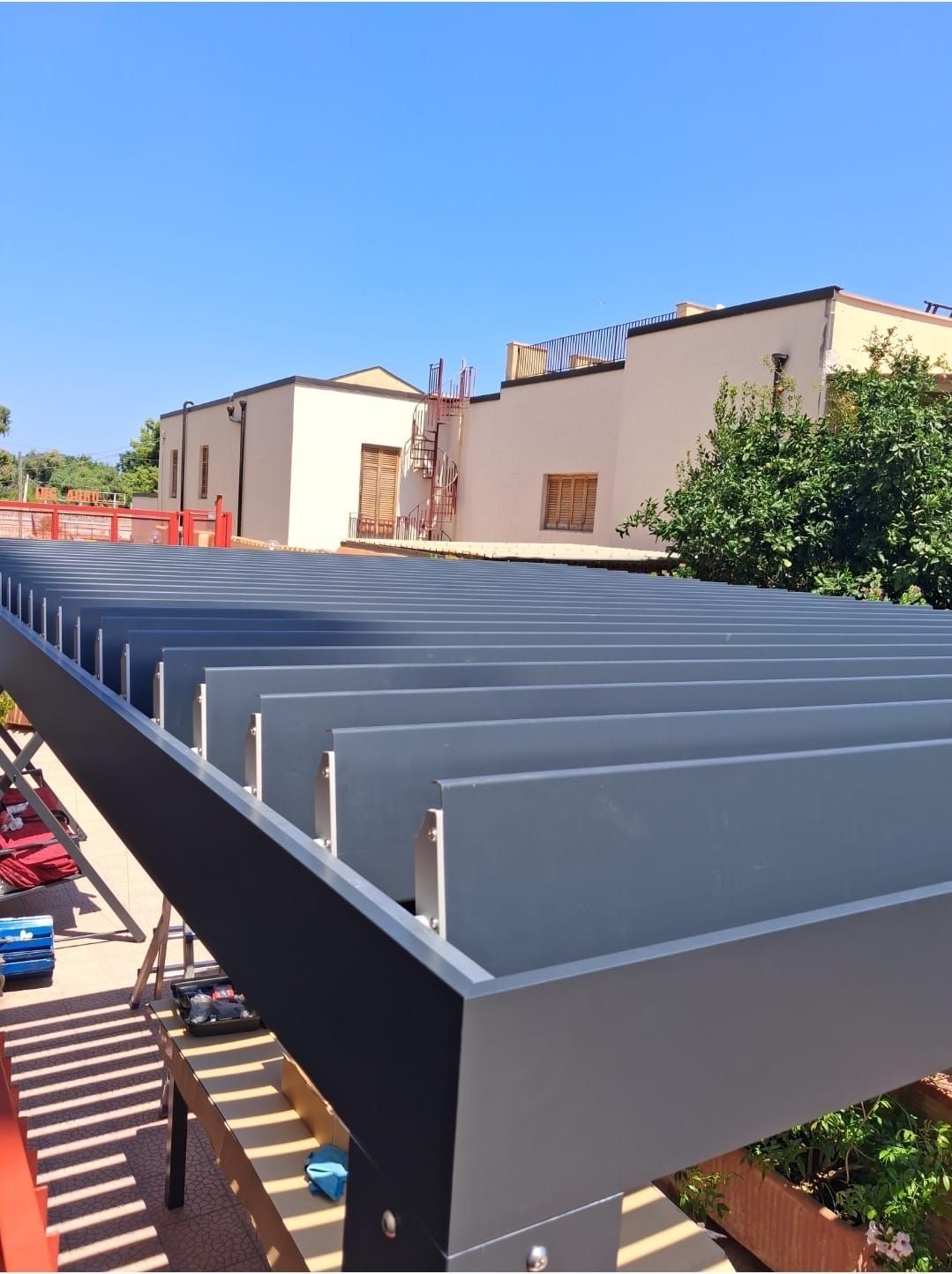 Pergola grigia e nera con doghe orizzontali, vista dal basso con edifici e cielo azzurro sullo sfondo.
