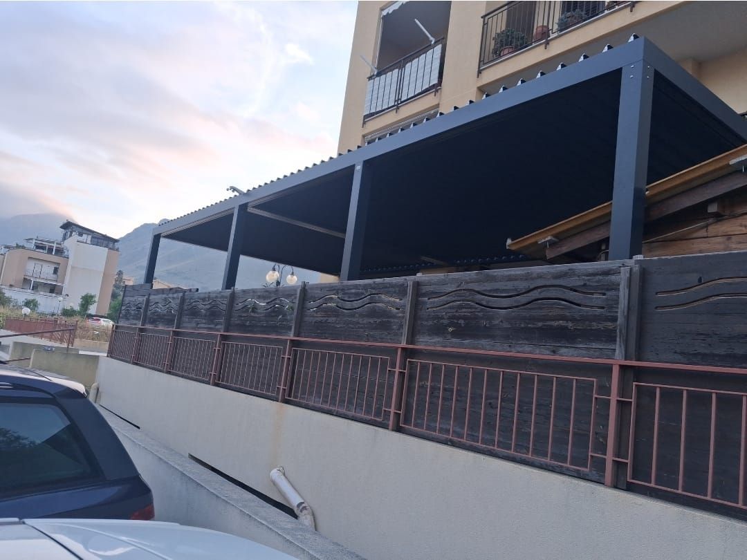 Pergola nera su un edificio, sopra una staccionata e una ringhiera in legno. Cielo grigio sullo sfondo.