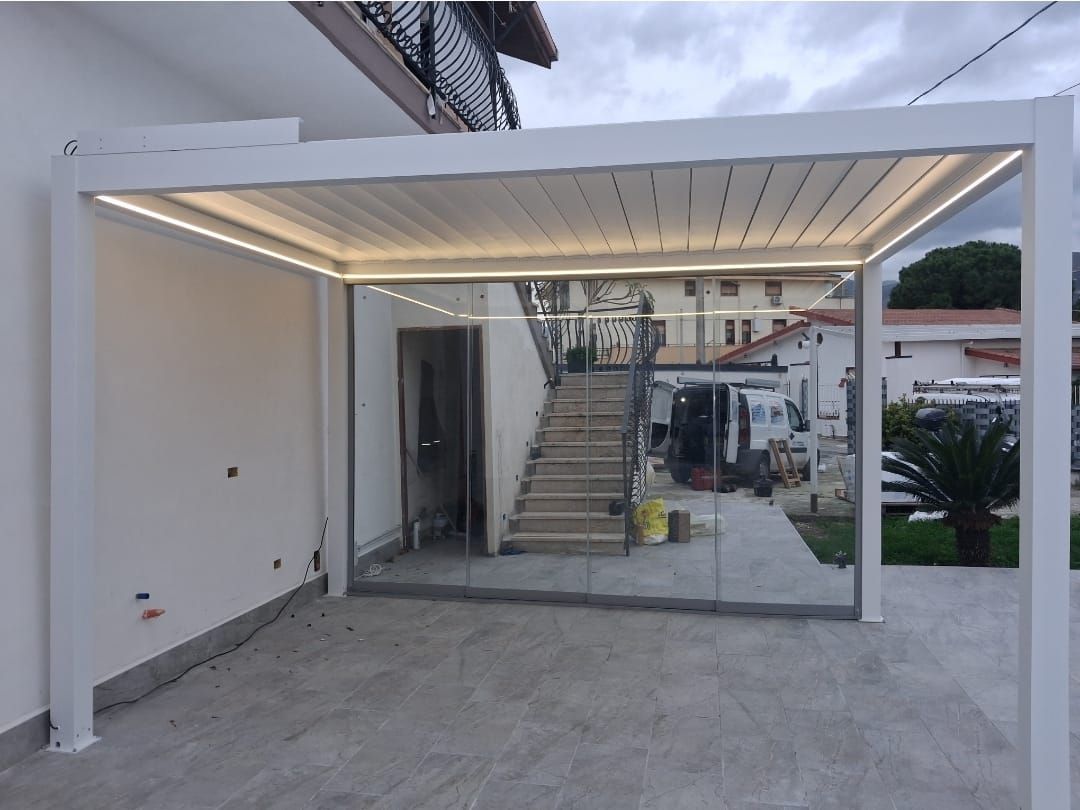 Pergola bianca con pareti in vetro e scale sullo sfondo, illuminata da luci zenitali.