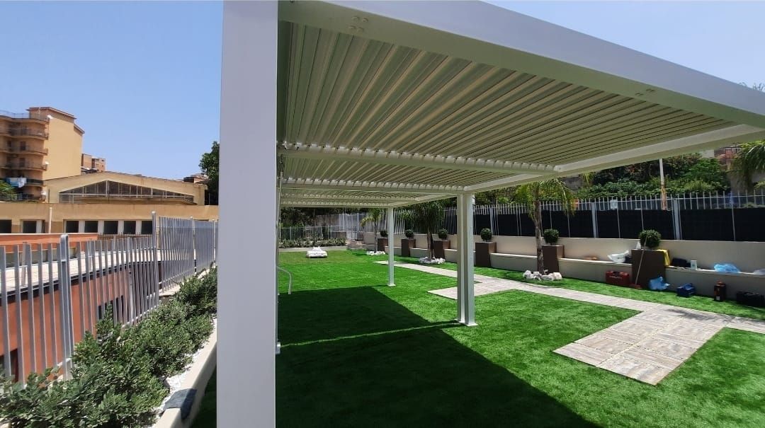 Pergola bianca su un prato verde con sedie a sdraio. Edificio sullo sfondo, persona seduta.