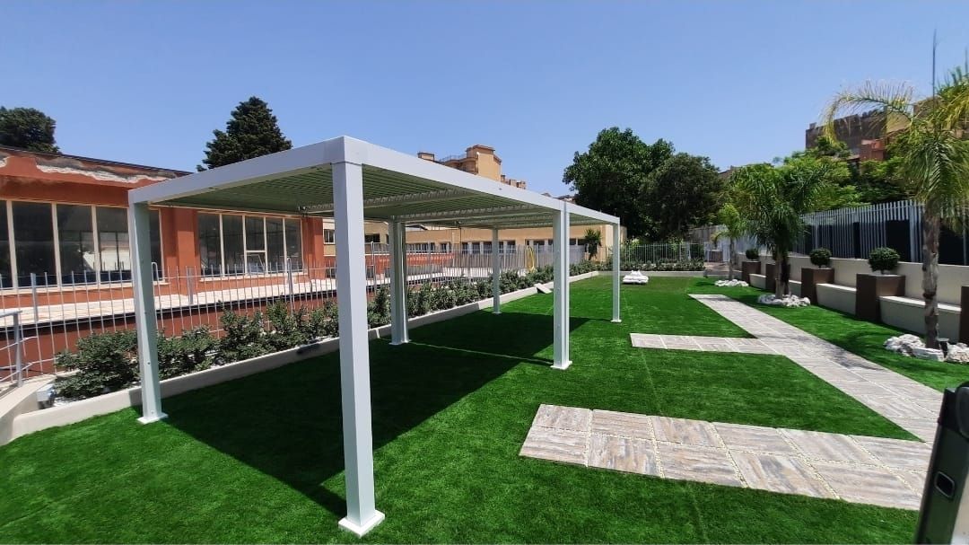 Un moderno pergolato con tetto a doghe si trova su un prato verde, proiettando ombre. In un ambiente esterno soleggiato.