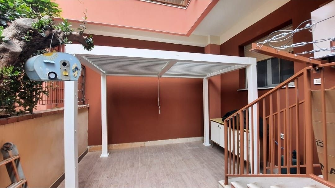 Pergola bianca sopra un patio con pareti rosso-marroni, una scala e un elettrodomestico attaccato.