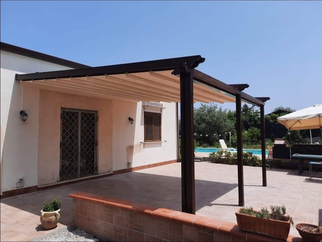 Pergola con tettoia beige retrattile che ombreggia il patio di un edificio chiaro, vicino a una piscina in una giornata di sole.