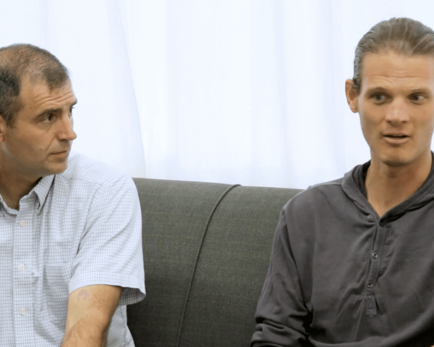 Two men sitting on a gray couch in a bright room, one speaking while the other listens.