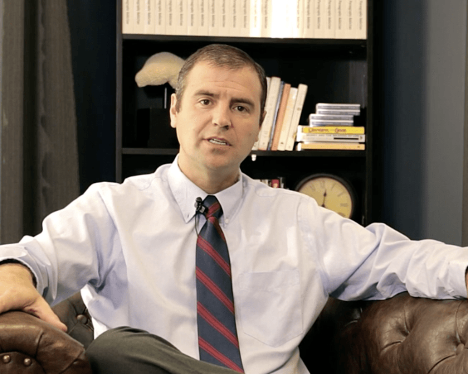 Man in white shirt and striped tie seated on a leather couch in an office, with bookshelves behind him