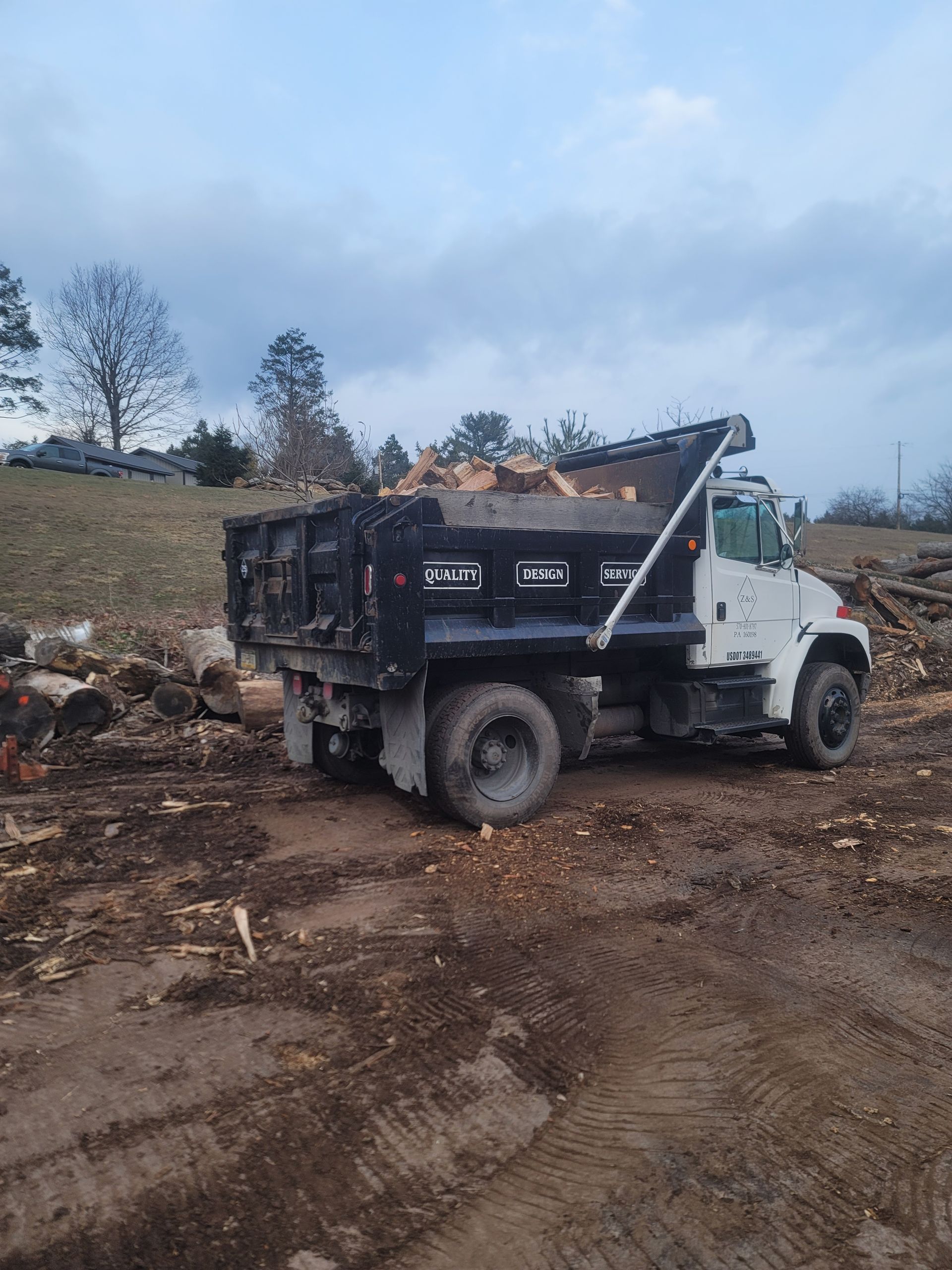Z&S Dump truck loaded with cut firewood parked on a muddy site with scattered logs and wood debris.  