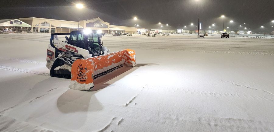 Skid steer with snow pusher clearing Hazleton parking lot at night.