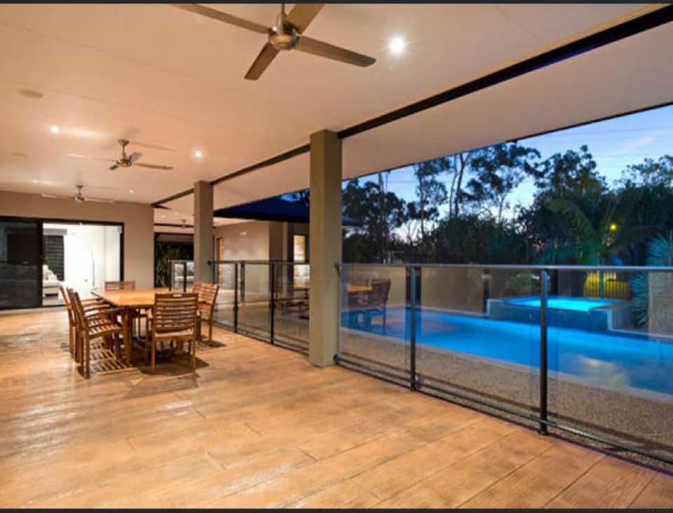 Patio with wooden furniture, overlooking a pool and trees. Ceiling fans and glass railings are visible — AKW Builders Pty Ltd In Leanyer, NT