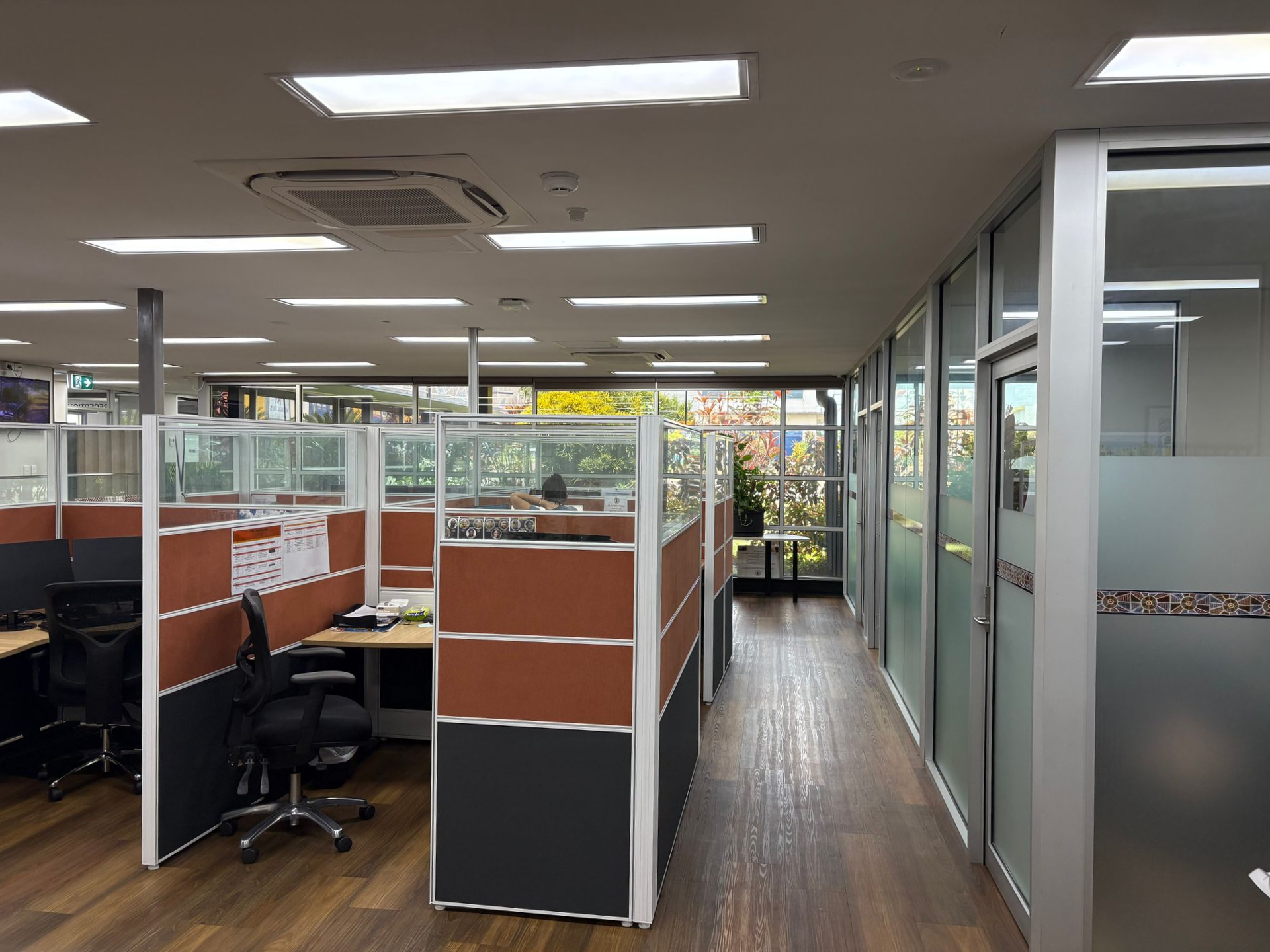 Office cubicles with brown and grey panels, and glass walls. Sunlight through windows.