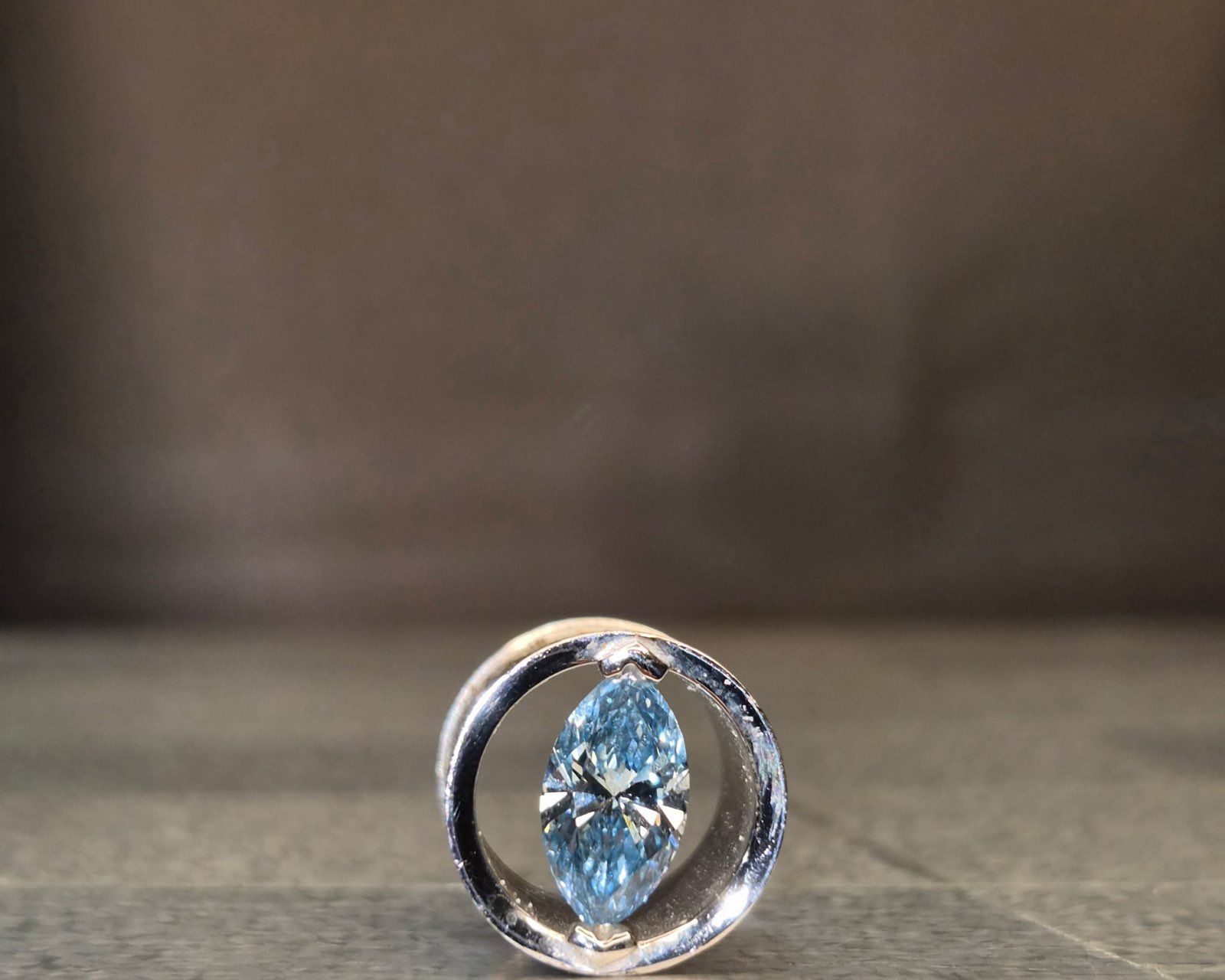 Light blue, marquise-cut gemstone set within a silver, ring-like holder; blurred background.