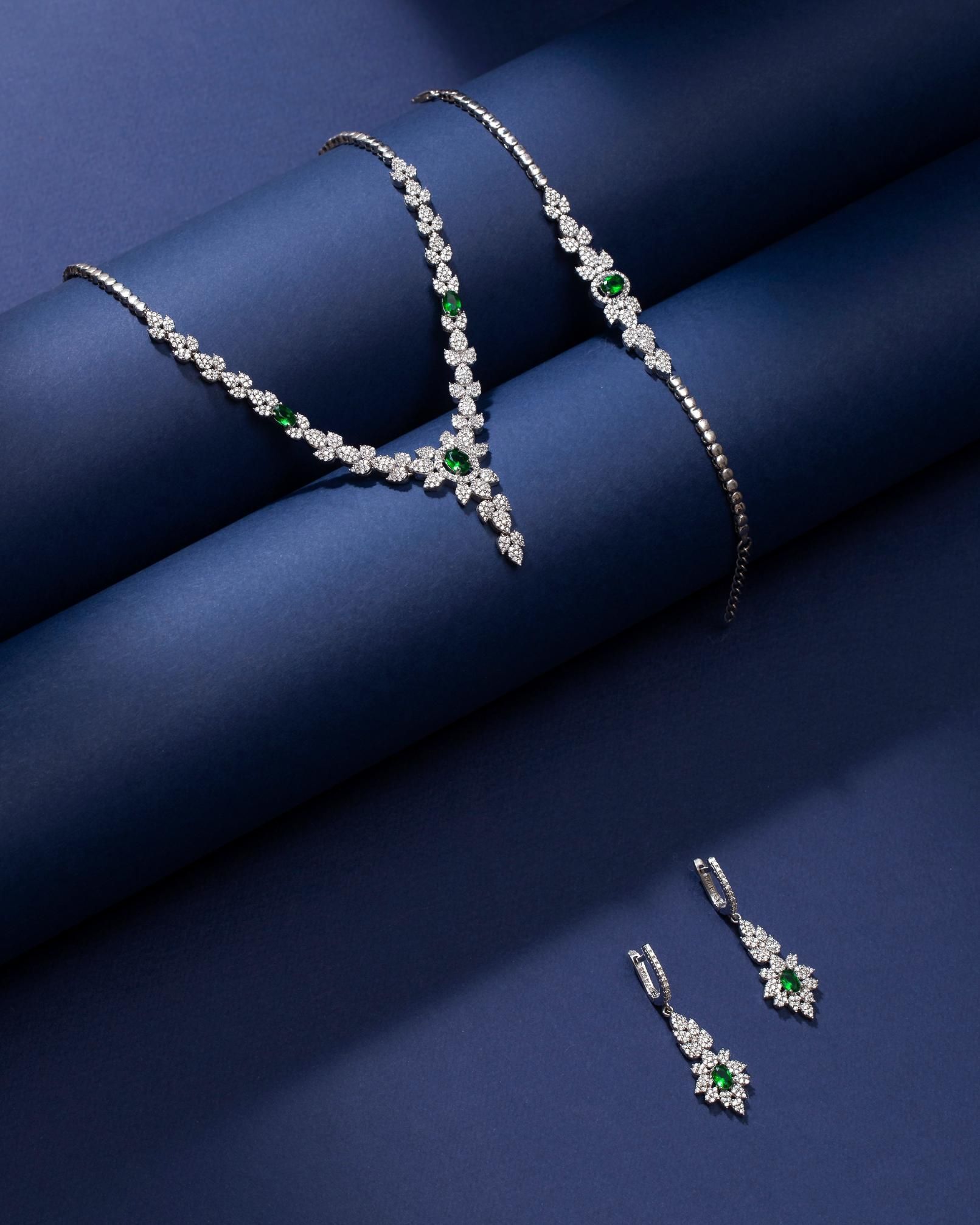 Jewelry set on blue background: necklace, bracelet, and earrings with emerald and diamond details.