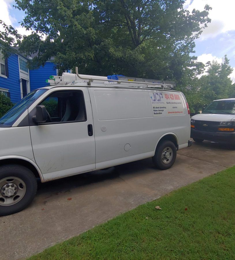 White service van parked on driveway next to dark car; green grass and blue house in the background.