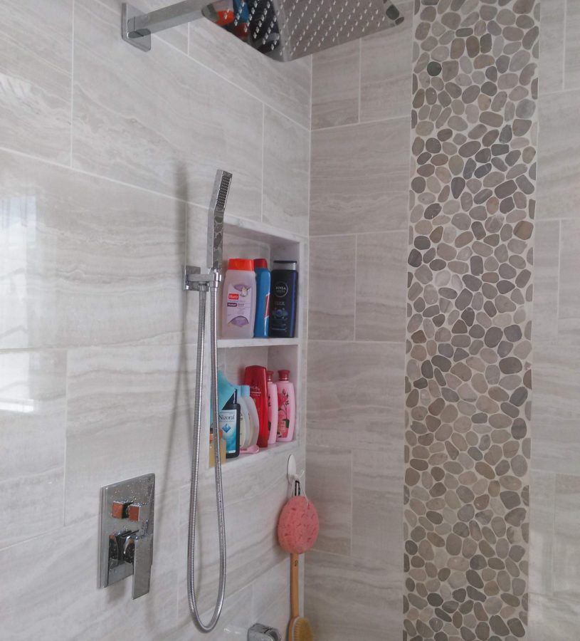 Shower with built-in shelf holding toiletries, alongside a pebble tile strip. Features rain showerhead and handheld wand.
