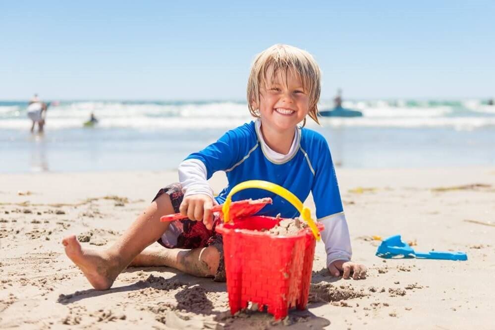 Boy with blonde hair smiles at the camera on a sandy beach, holding a red bucket filled with sand and toys.