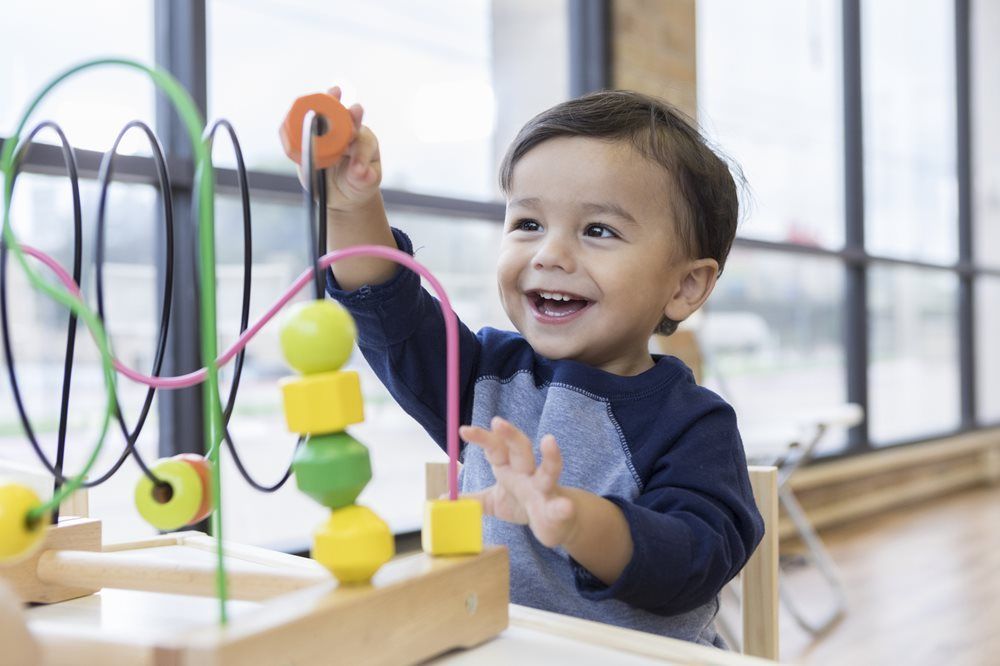 Smiling child playing with a wooden bead maze, near a window.