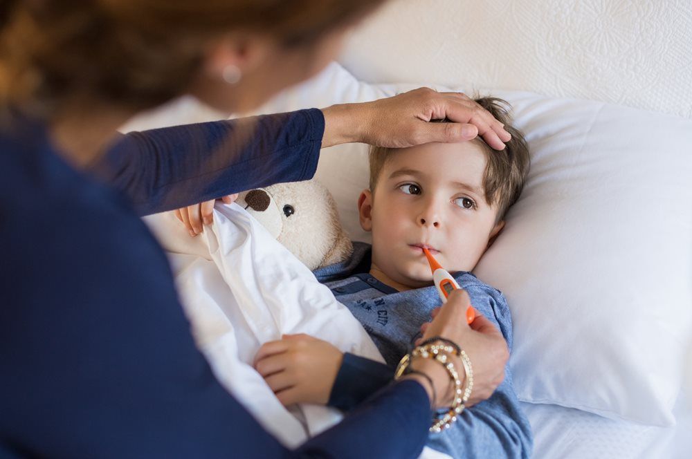 Woman checking a child's temperature with a thermometer while touching his forehead. The child is in bed, looking unwell.
