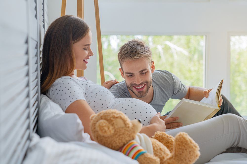 Pregnant woman and man in bed reading a book, smiling. Teddy bear on the bed.