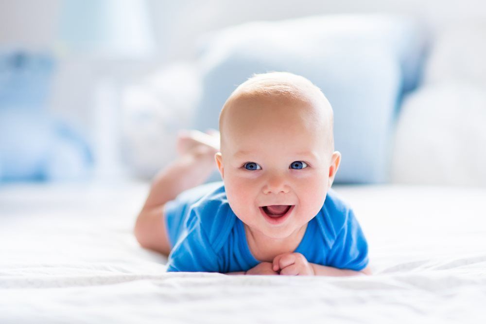 Smiling baby wearing blue on a white surface, with blue pillows in the background.