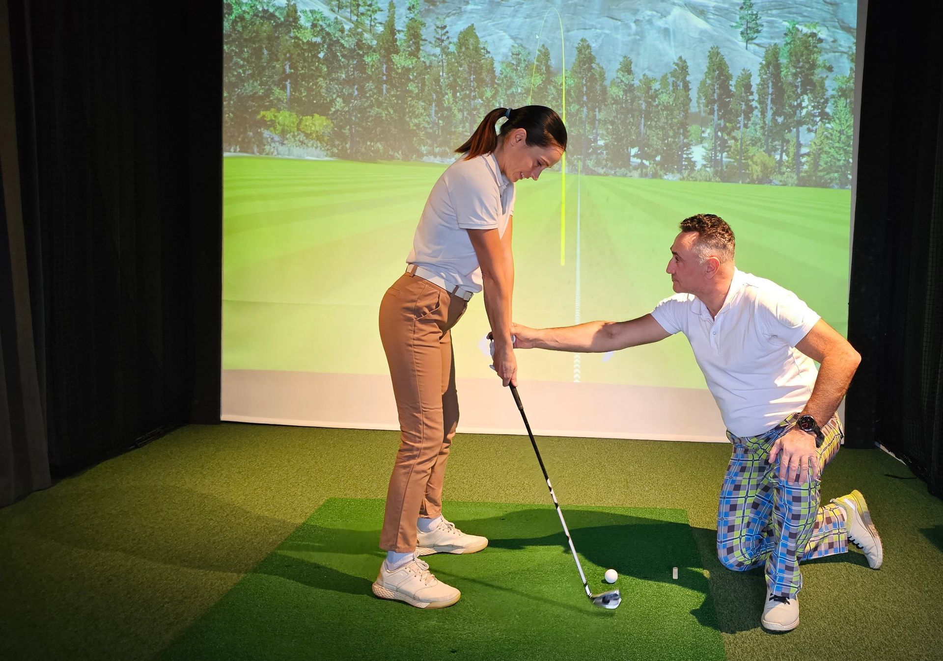 Two people practicing golf indoors, one kneeling to help the other hold a club on a green turf mat.