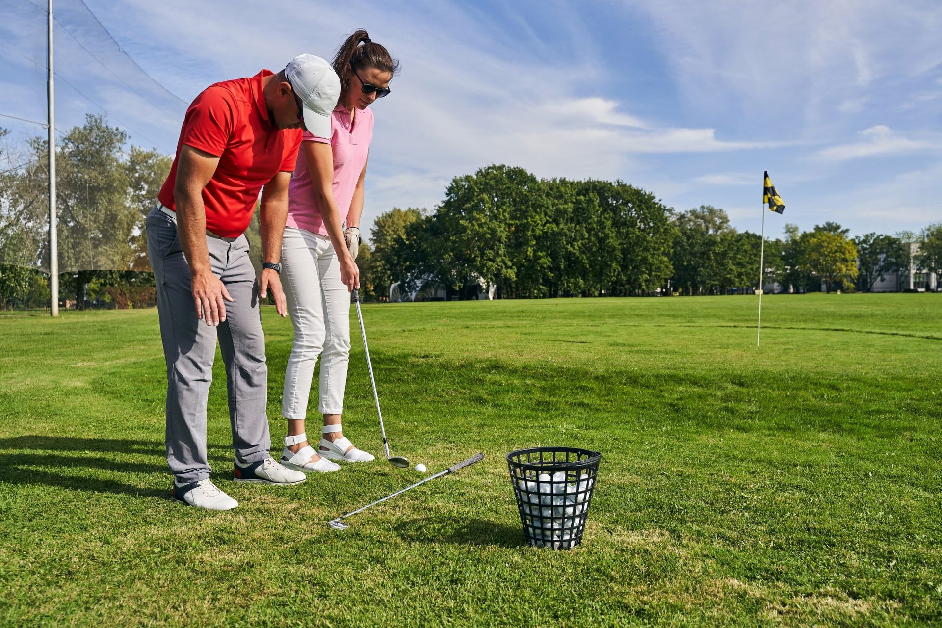 Two golfers practicing putting on a green beside a ball basket and flagstick