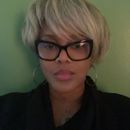 Woman with blonde pixie cut, wearing glasses and black top, posing in front of a green wall.