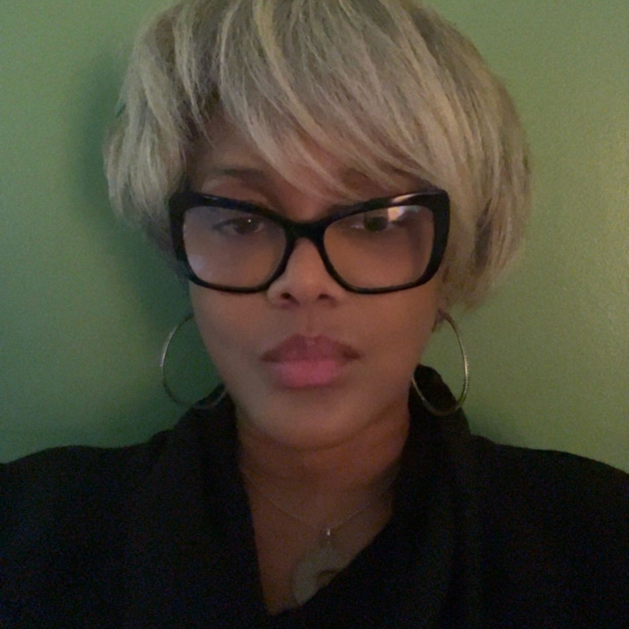 Woman with short blonde hair, glasses, and hoop earrings; wearing black; against a green backdrop.
