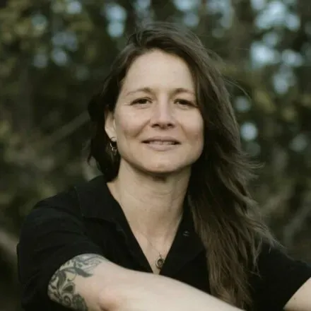 Woman with long hair, smiling, wearing a black shirt, and has a tattoo on her arm, set outdoors.