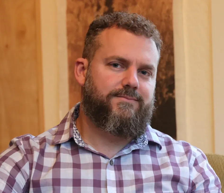 Man with beard and plaid shirt, seated with a slight smile, indoors.