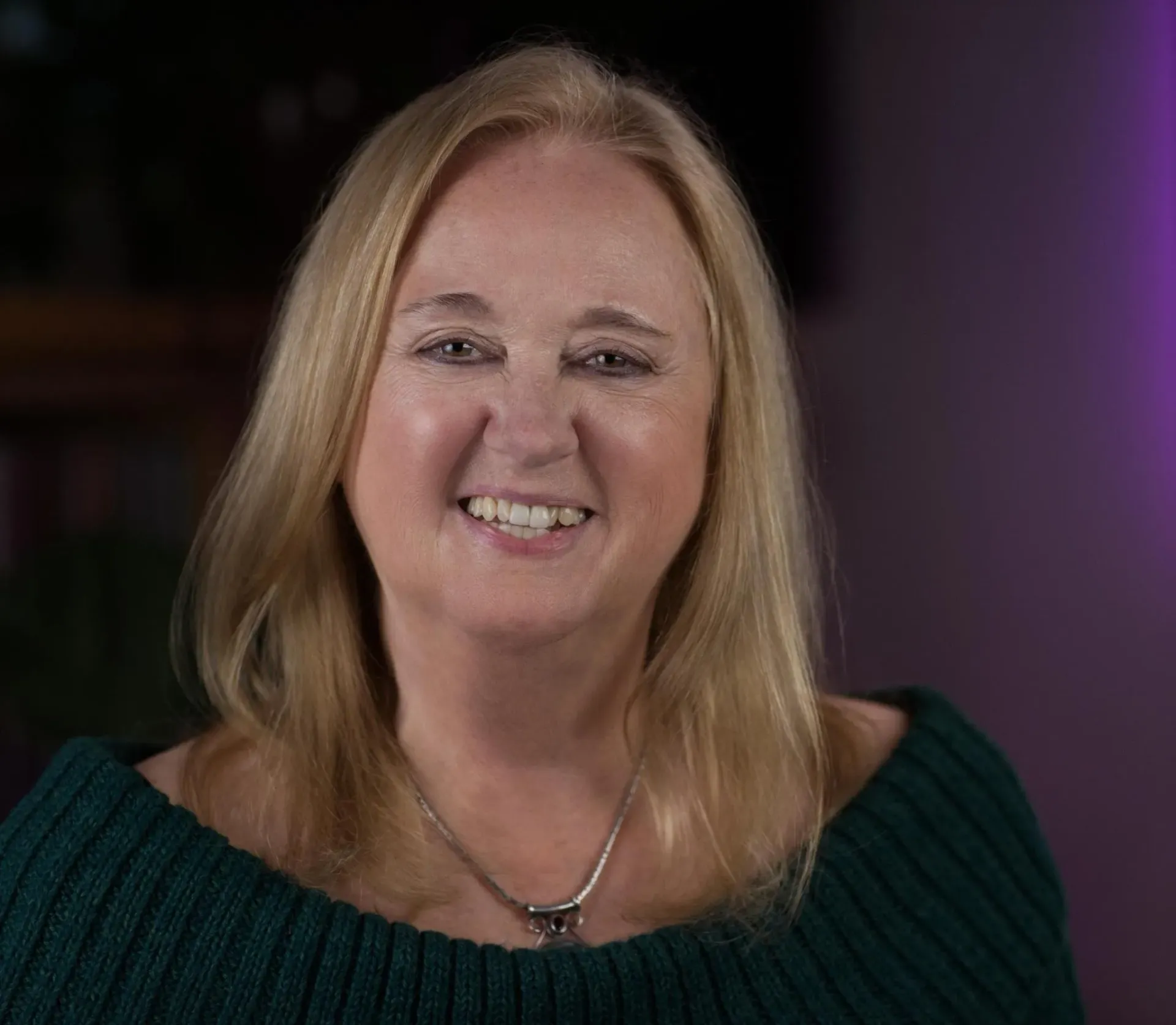 Woman with blonde hair smiles, wearing a green sweater and a necklace, indoors.