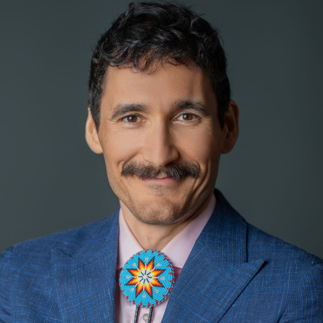 Man with mustache wearing bolo tie, suit, and pink shirt smiles against a dark gray background.