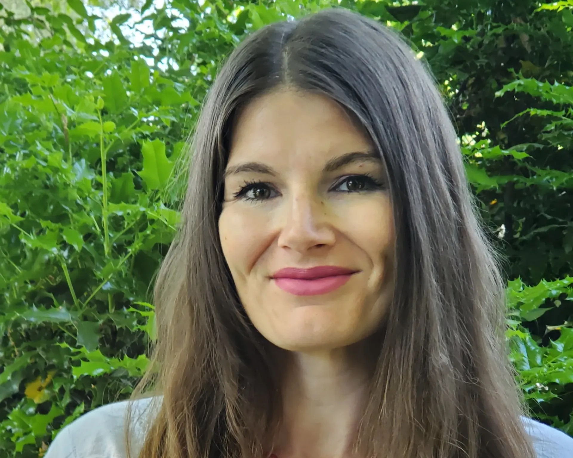 Woman with long brown hair smiles in front of green foliage.