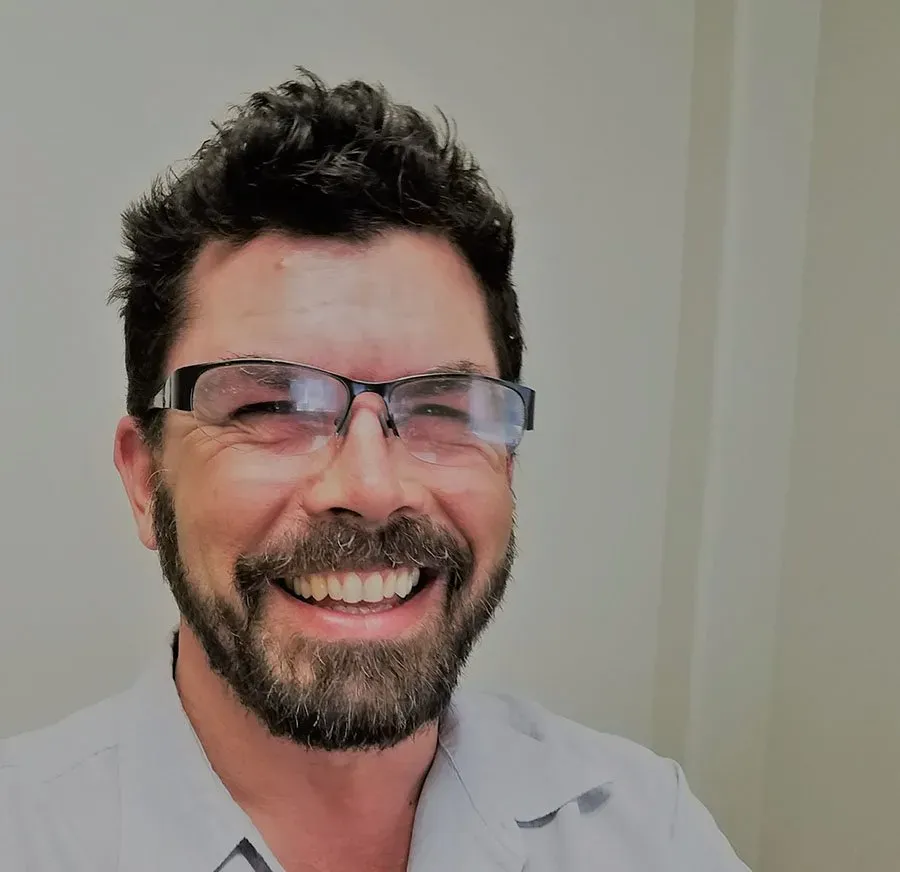 Man with beard and glasses smiling, light-colored shirt, neutral background.