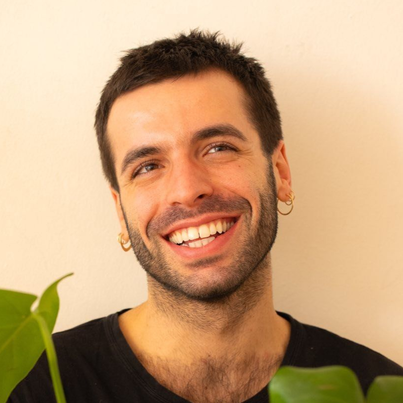 Smiling man with dark hair, gold hoop earrings, and a black shirt, holding a monstera plant.