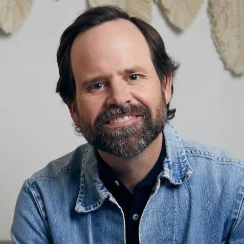 Smiling man with dark hair and beard in a denim jacket. Neutral setting with decorative wall art.