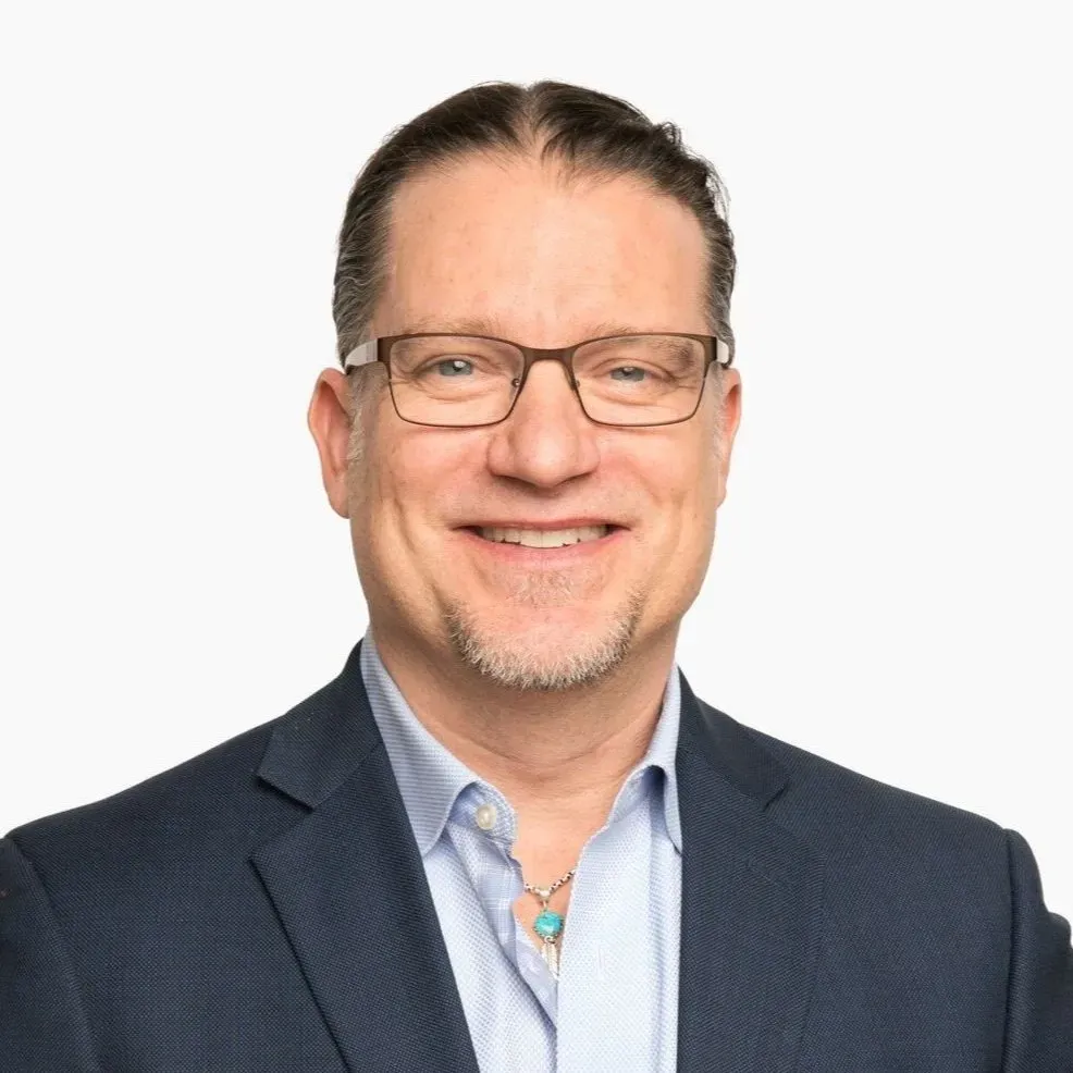 Man with glasses smiles, wearing blue jacket and light blue shirt.