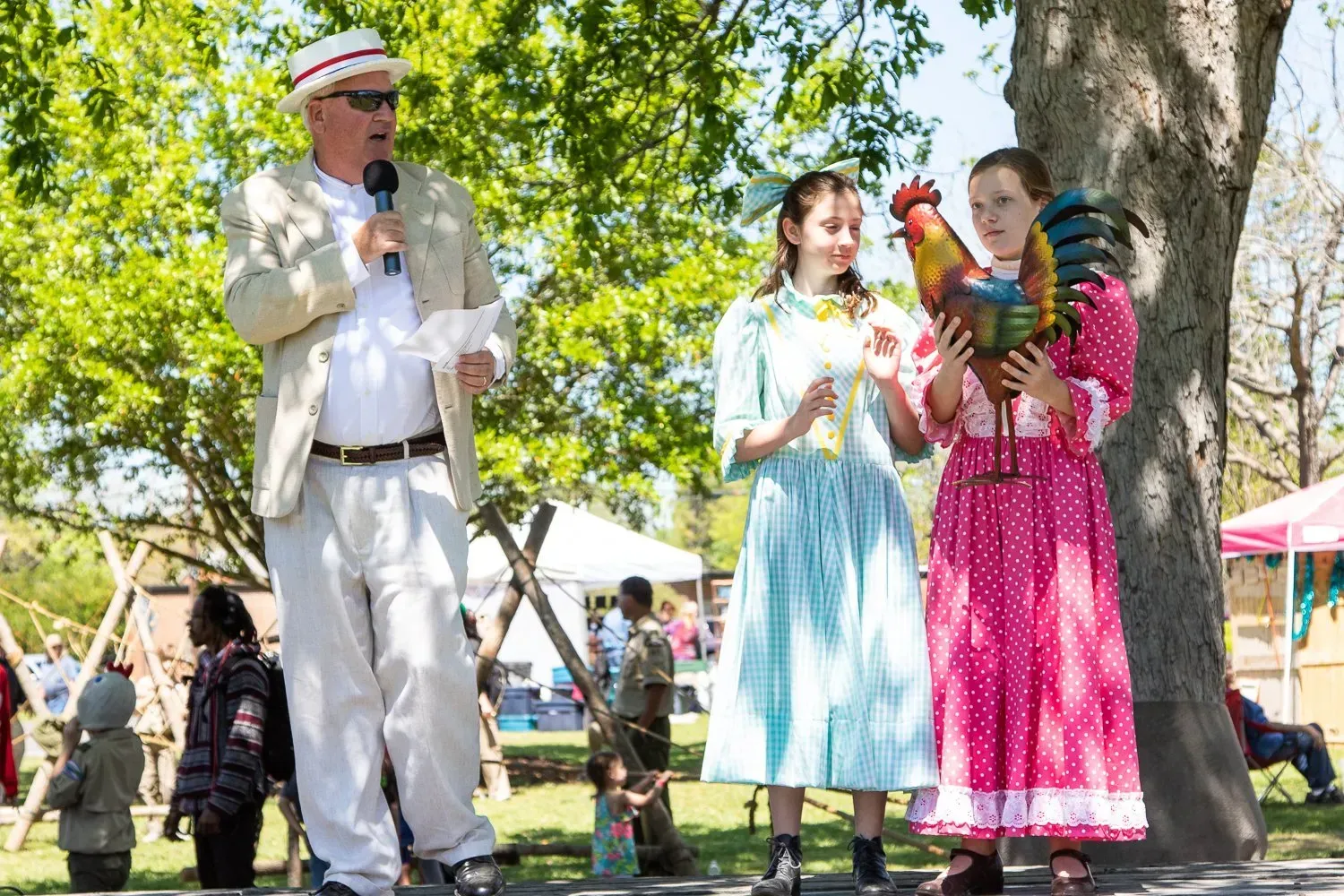 A man and two girls are standing next to each other in a park holding a rooster.
