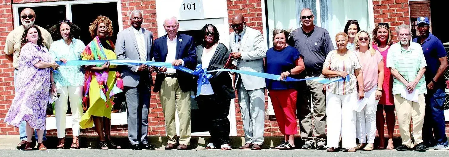 A group of people are standing in front of a brick building cutting a blue ribbon.