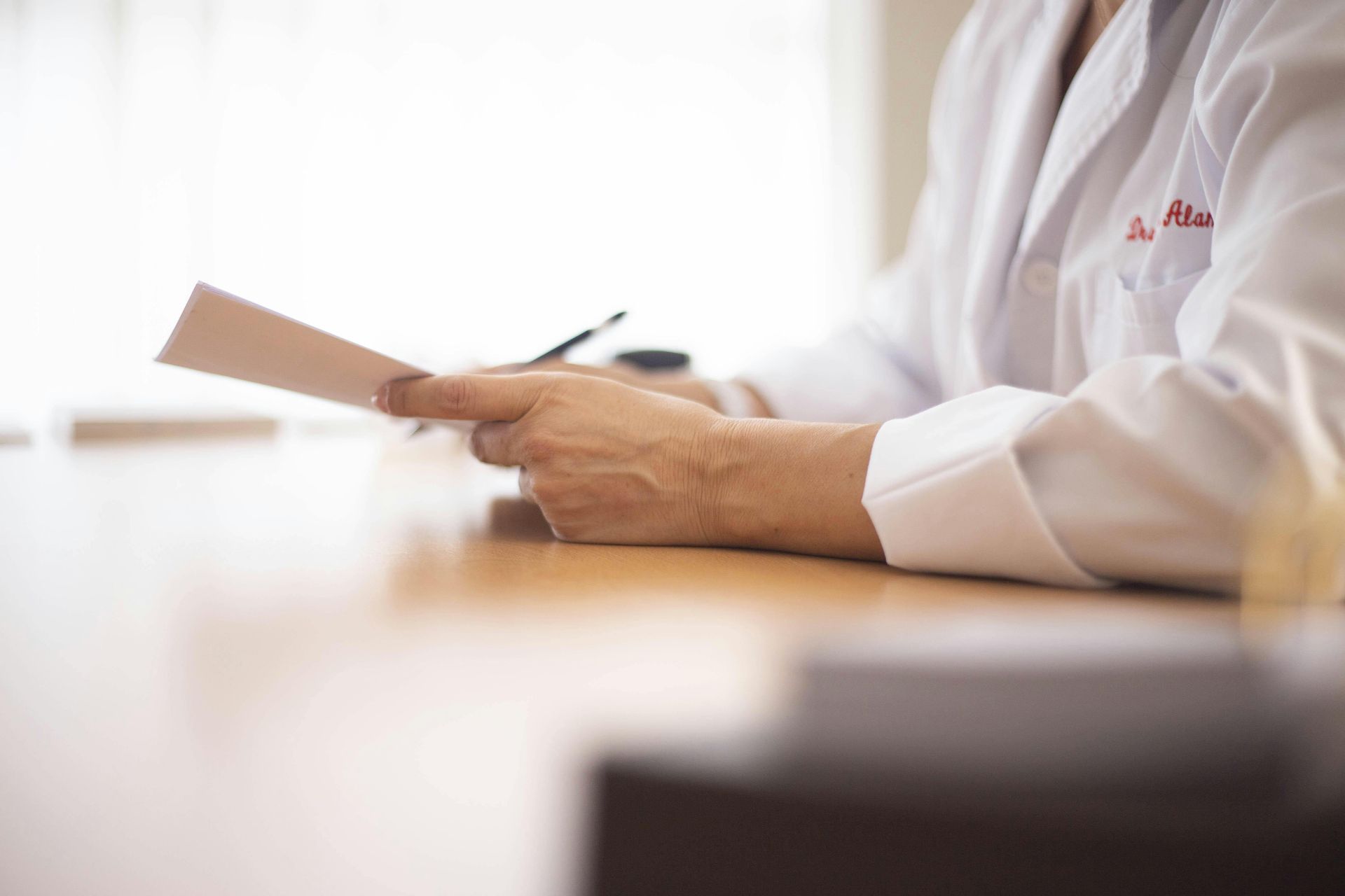 Doctor in white coat, holding papers, sitting at a wooden table, indoors.